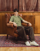 A young man sits in a brown leather armchair wearing an olive green t-shirt, dark brown pants, and white sneakers, set against a background of wood paneling and a patterned rug.