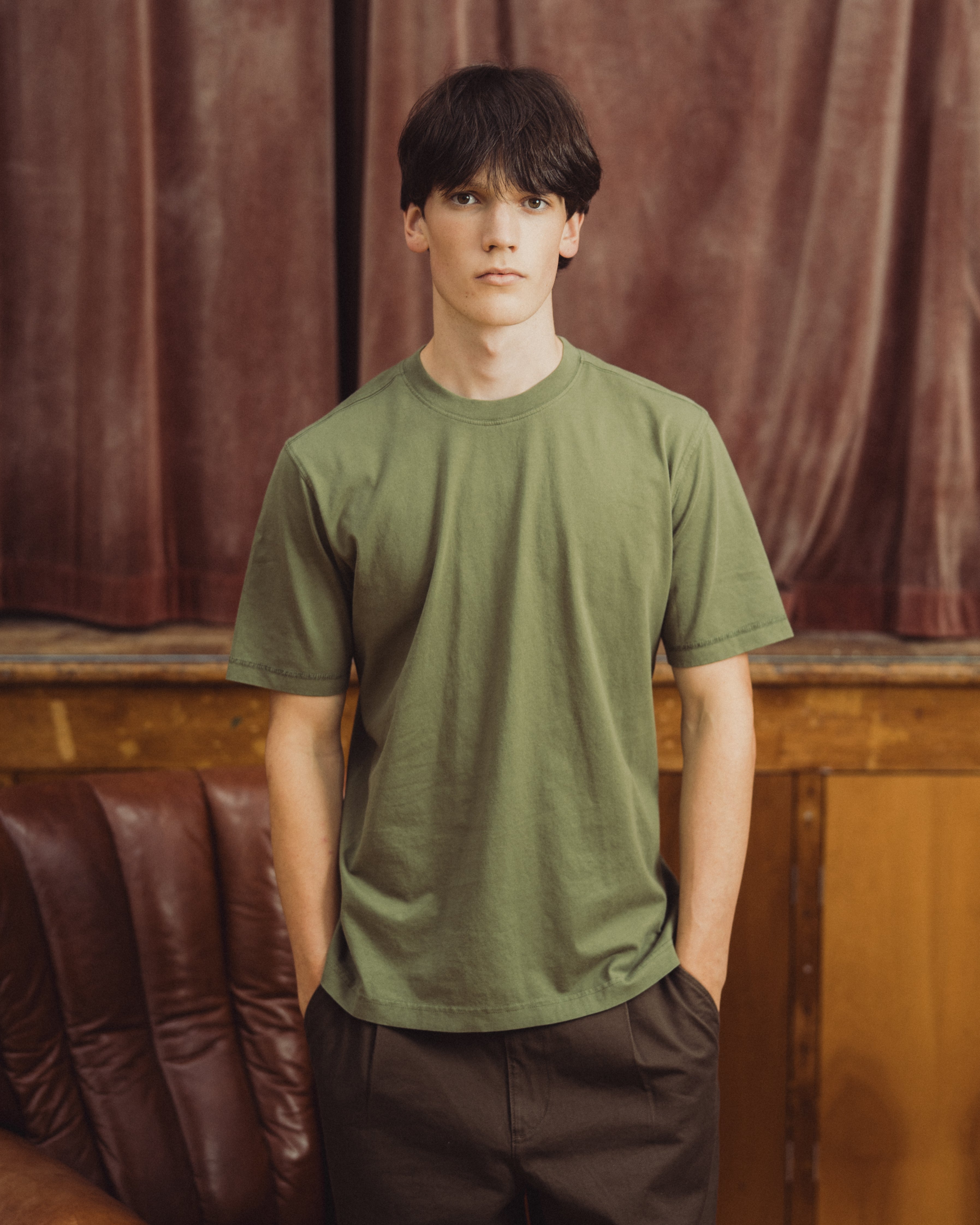 A young man wearing a muted olive green crewneck t-shirt and dark brown trousers, standing in front of a brown leather chair and a brown velvet curtain.