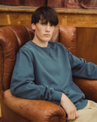 A young man with dark hair sits in a brown leather armchair, wearing a muted slate blue crewneck sweatshirt and light beige trousers.