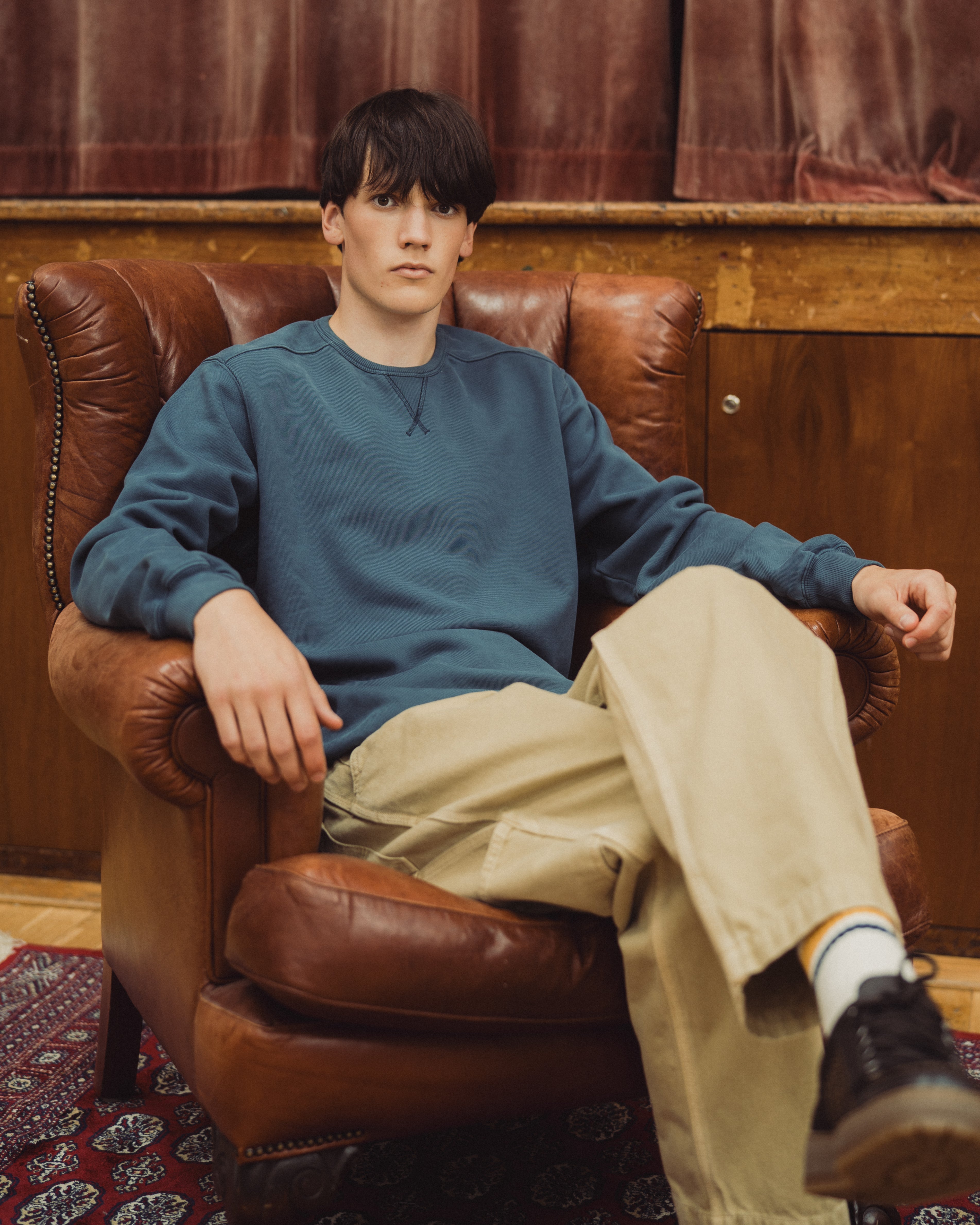 A young man sits in a brown leather armchair wearing a muted blue crewneck sweatshirt, beige cargo pants, white socks, and black sneakers in a warm, wood-paneled room.
