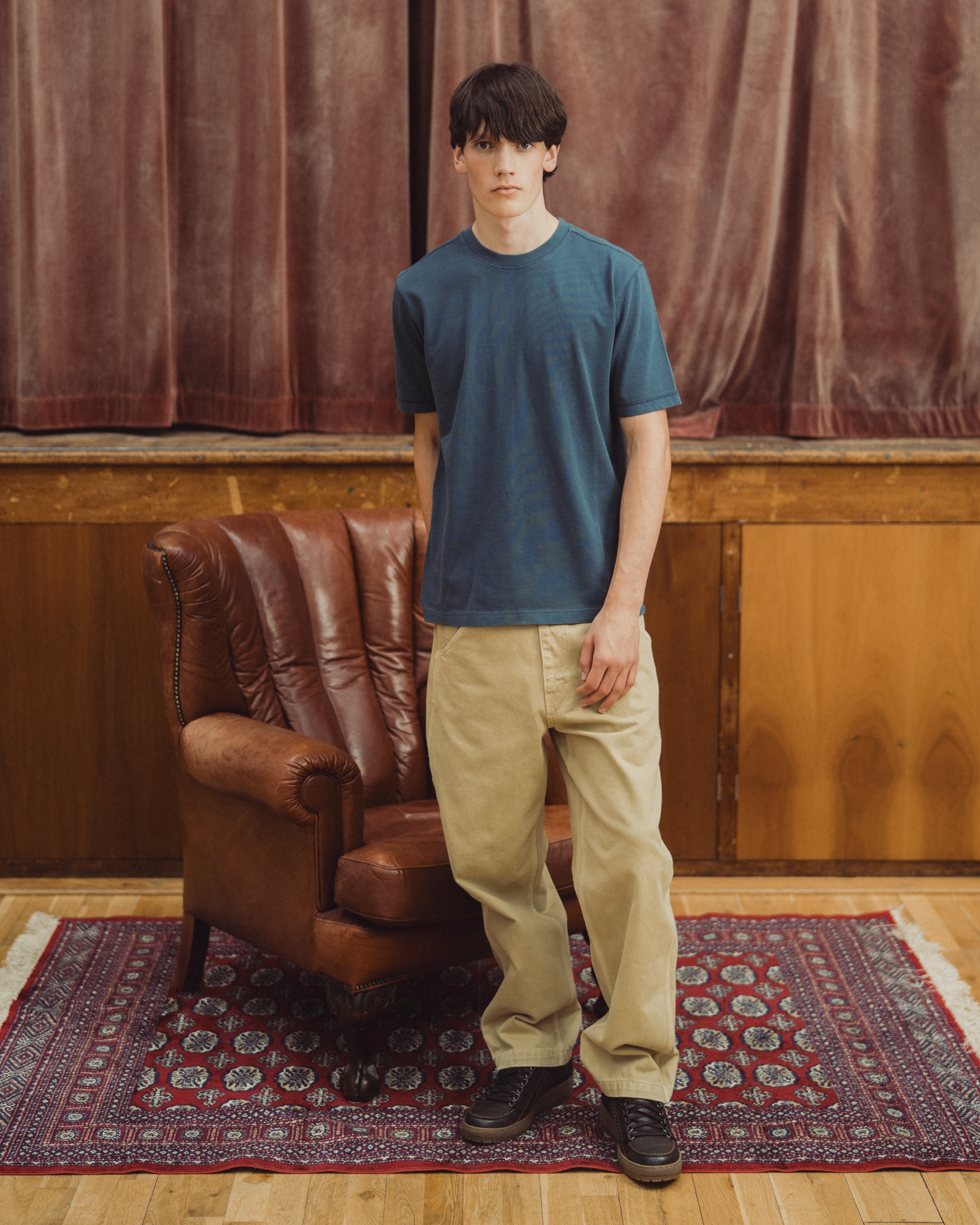 A man stands in a room wearing a muted blue crew-neck t-shirt, beige pants, and dark shoes, positioned next to a brown leather armchair on a patterned rug.