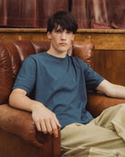 A young man with dark hair sits in a brown leather armchair, wearing a muted slate blue crewneck t-shirt and beige cargo pants.