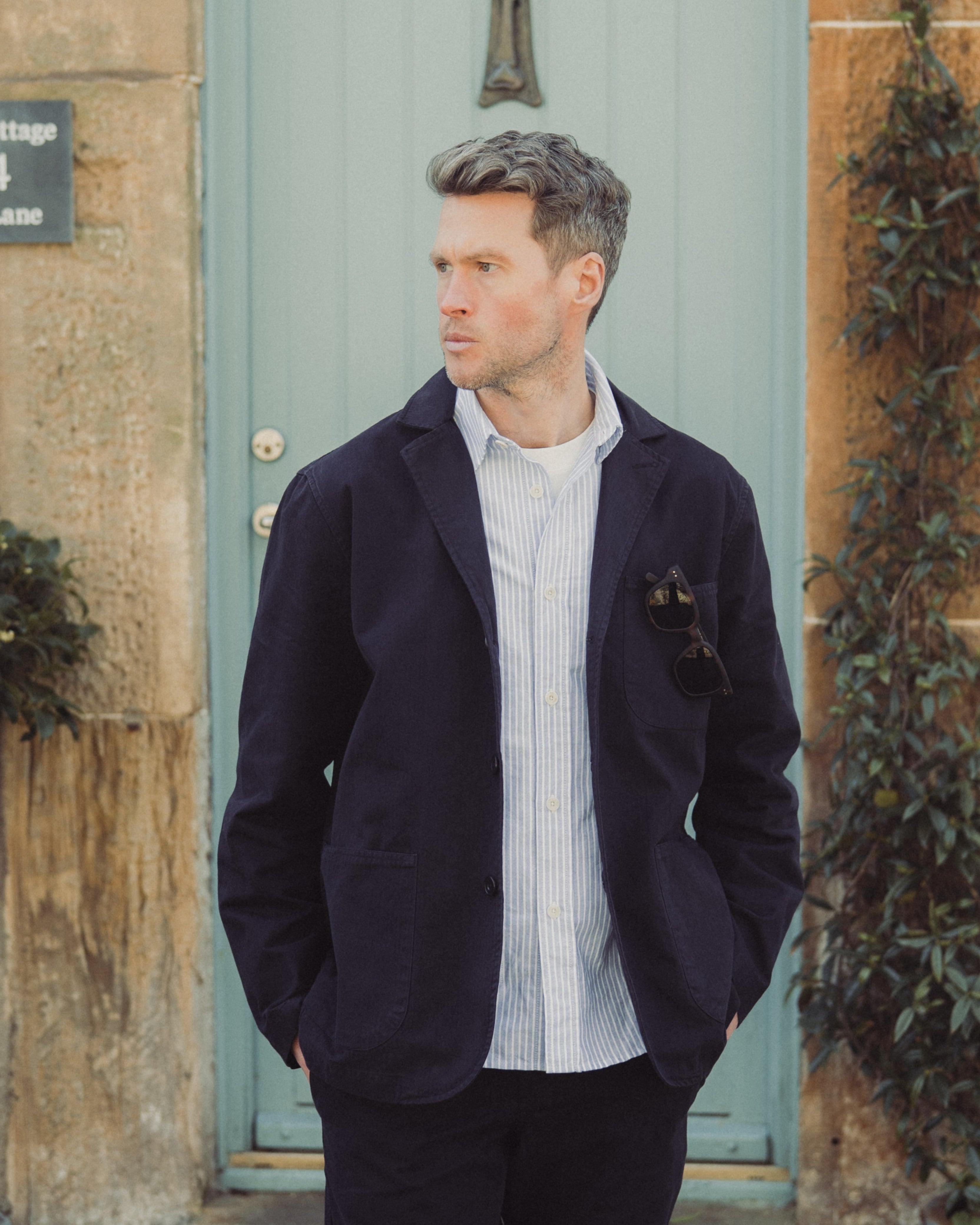 A man wearing a dark navy blazer over a white striped shirt and dark trousers, standing in front of a light blue cottage door with stone walls and potted plants.