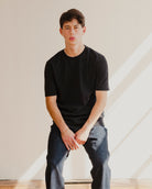 Person wearing a black t-shirt and gray pants sitting against a neutral background