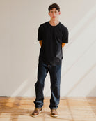 Man wearing a black t-shirt and blue jeans standing against a plain wall.