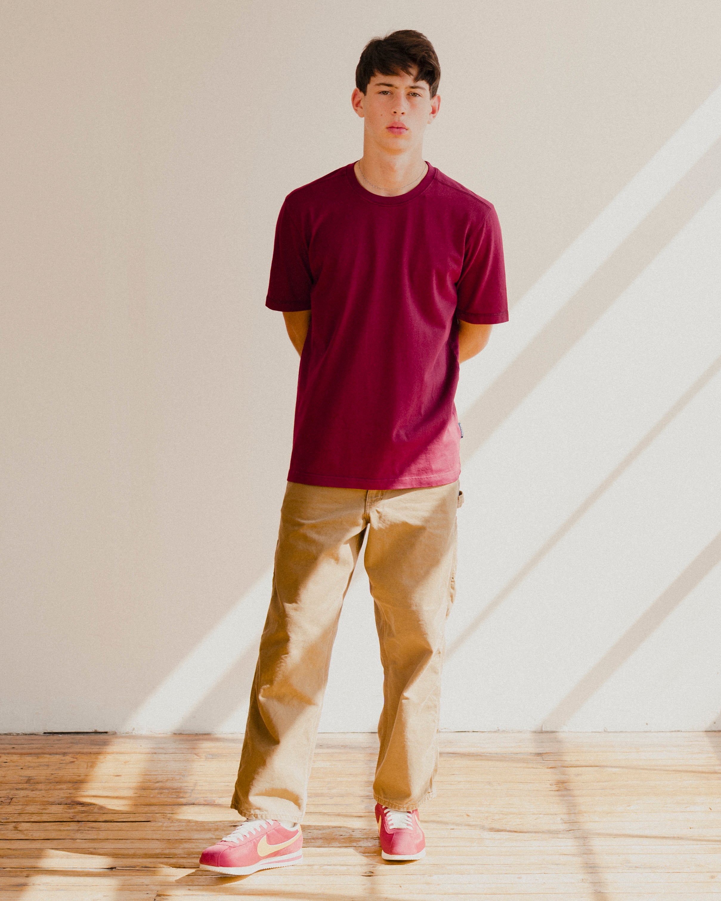 Man wearing a maroon red t-shirt and beige pants standing in a room with sunlight streaming through a window.