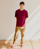 Man wearing a maroon red t-shirt and beige pants standing in a room with sunlight streaming through a window.