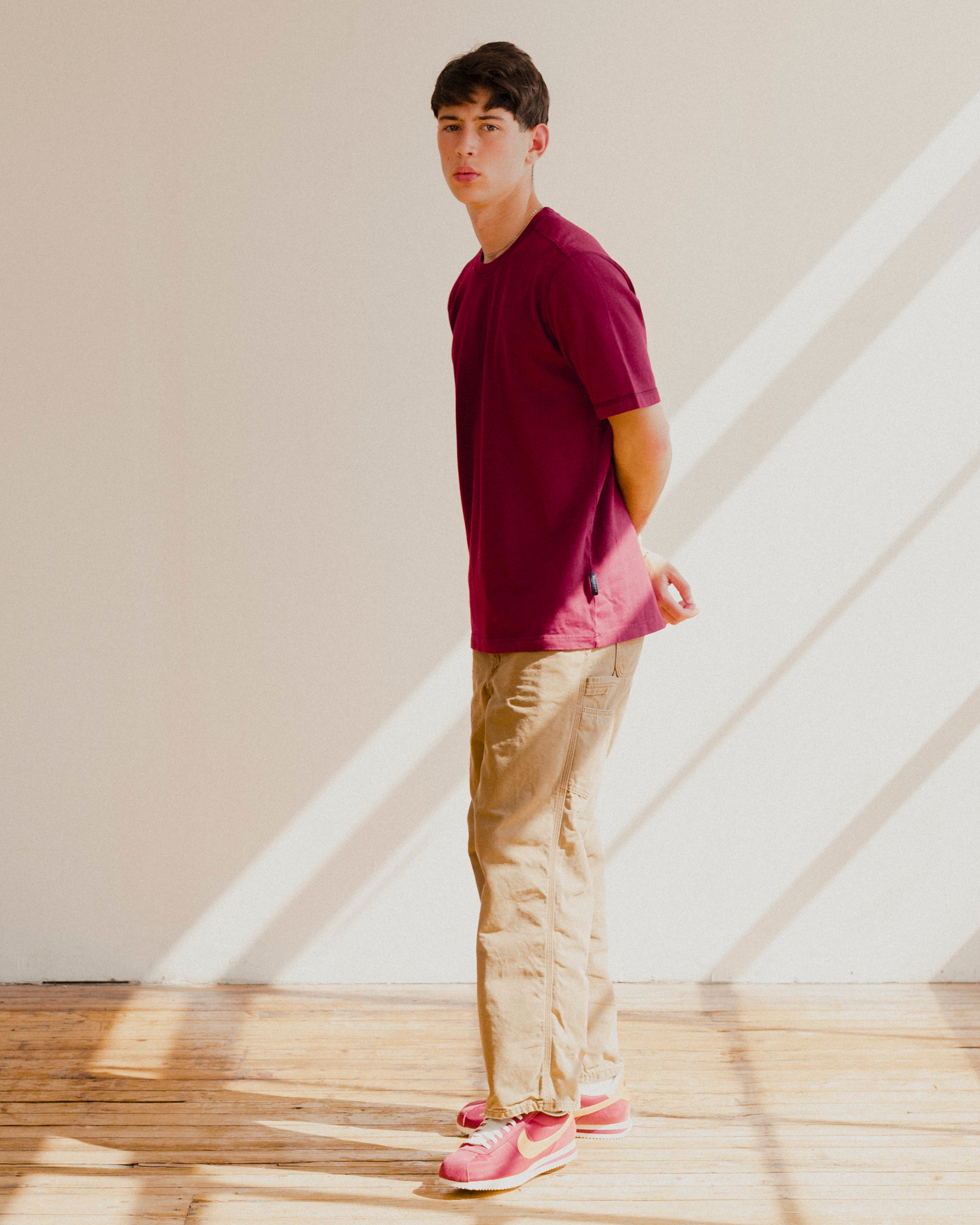 Man wearing a maroon red shirt and beige pants standing in a room with wooden floor and white walls.