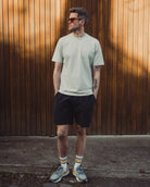 Man wearing a light green t-shirt and black shorts standing against a wooden wall.