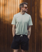 Man wearing a light green t-shirt and black shorts standing against a wooden wall.