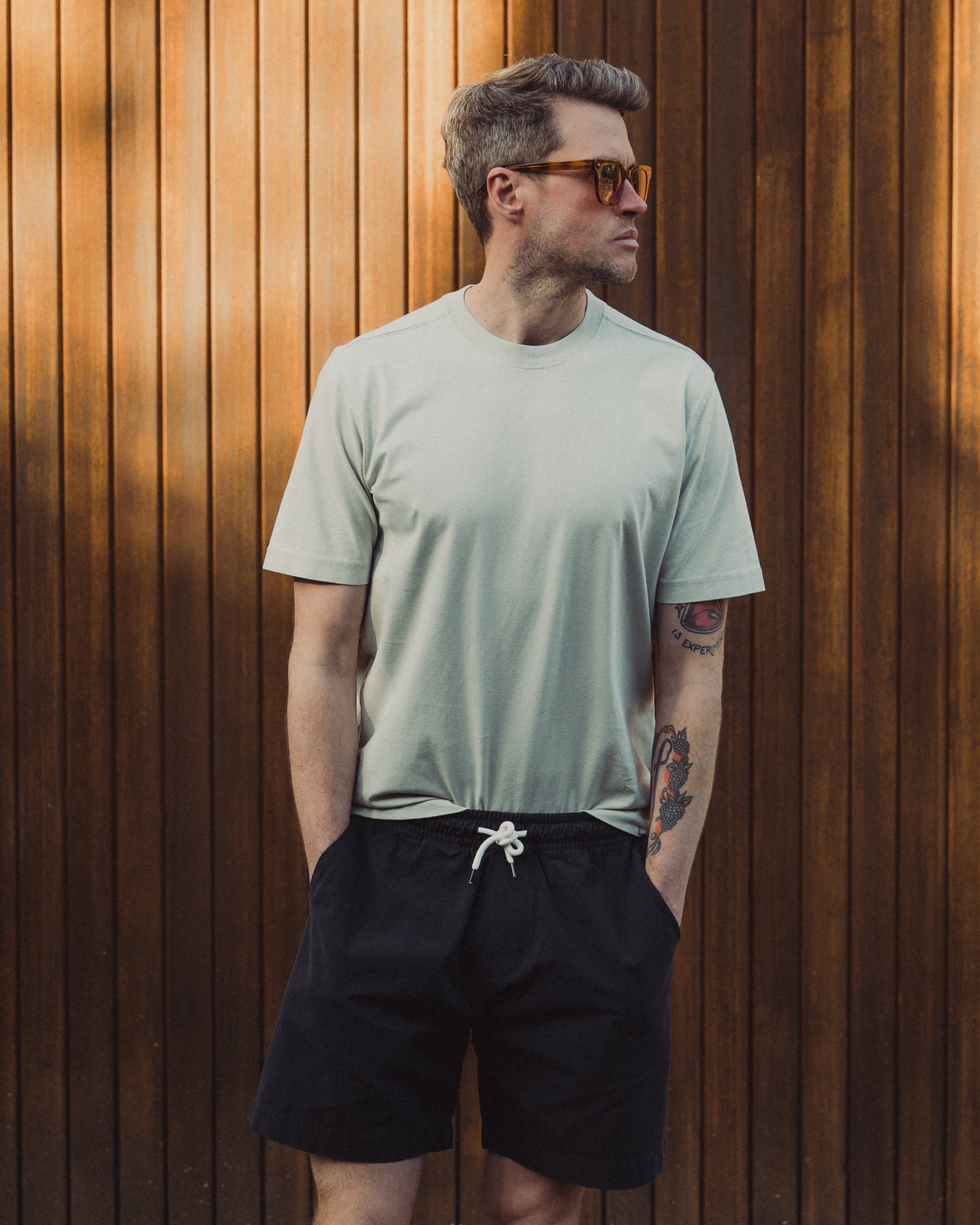 Man wearing a light green t-shirt and black shorts standing against a wooden wall.