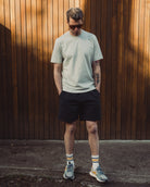 Man wearing a light green t-shirt and black shorts standing against a wooden wall.