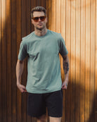 Man wearing a light green t-shirt and black shorts standing against a wooden wall.