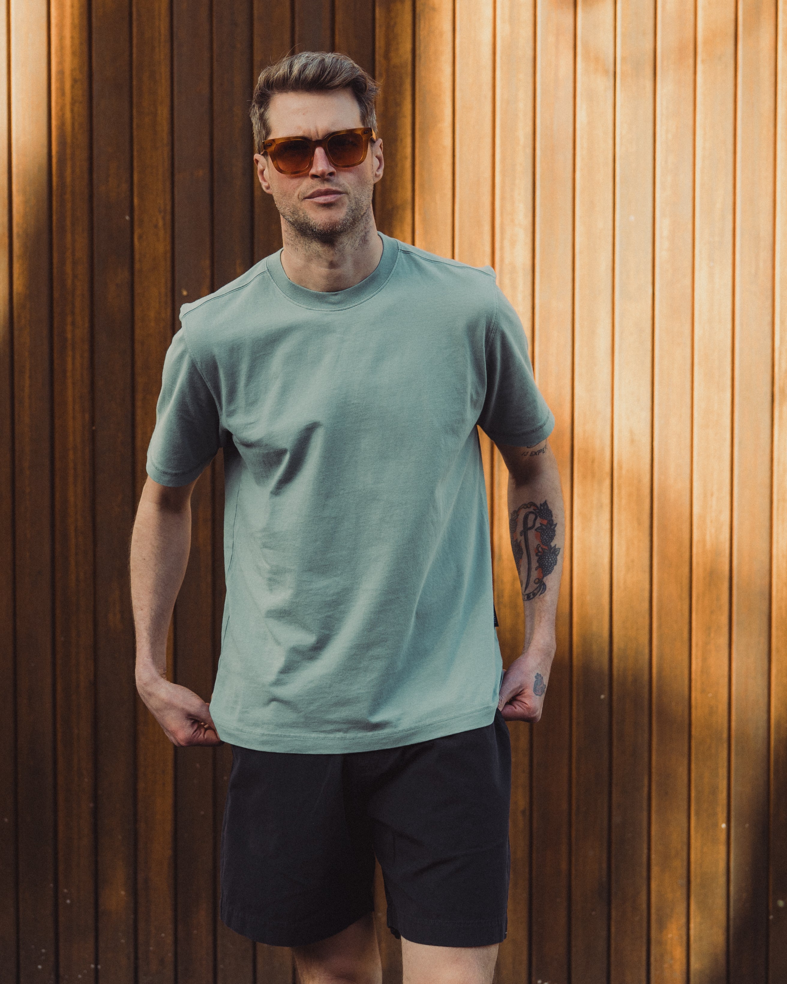 Man wearing a light green t-shirt and black shorts standing against a wooden wall.