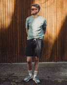Man wearing sunglasses, a light gray t-shirt, black shorts, and colorful socks standing against a wooden wall.
