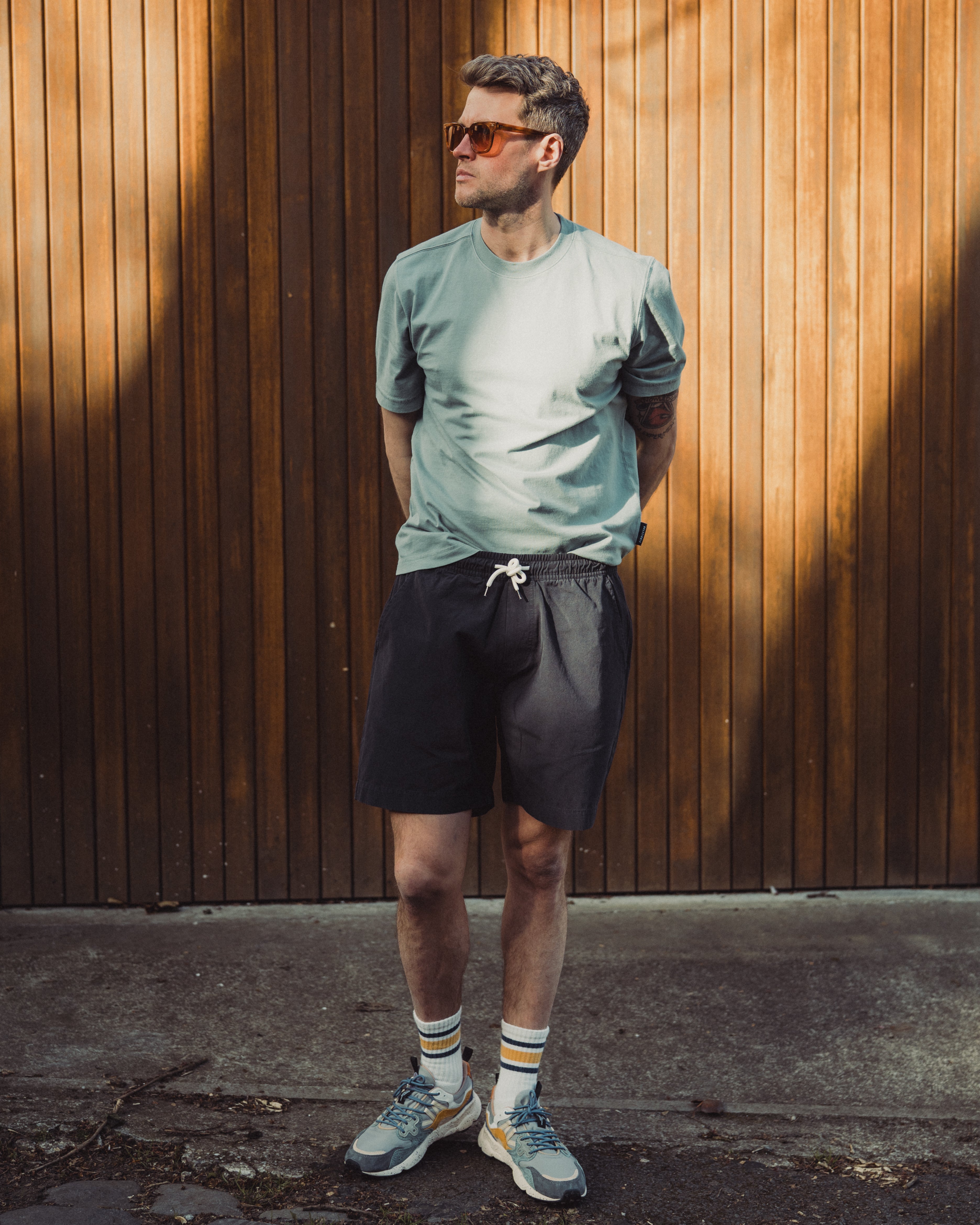 Man wearing sunglasses, a light gray t-shirt, black shorts, and colorful socks standing against a wooden wall.