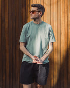 Man wearing a green t-shirt and black shorts standing against a wooden wall.