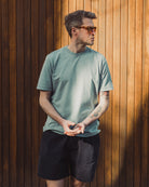 Man wearing a green t-shirt and black shorts standing against a wooden wall.