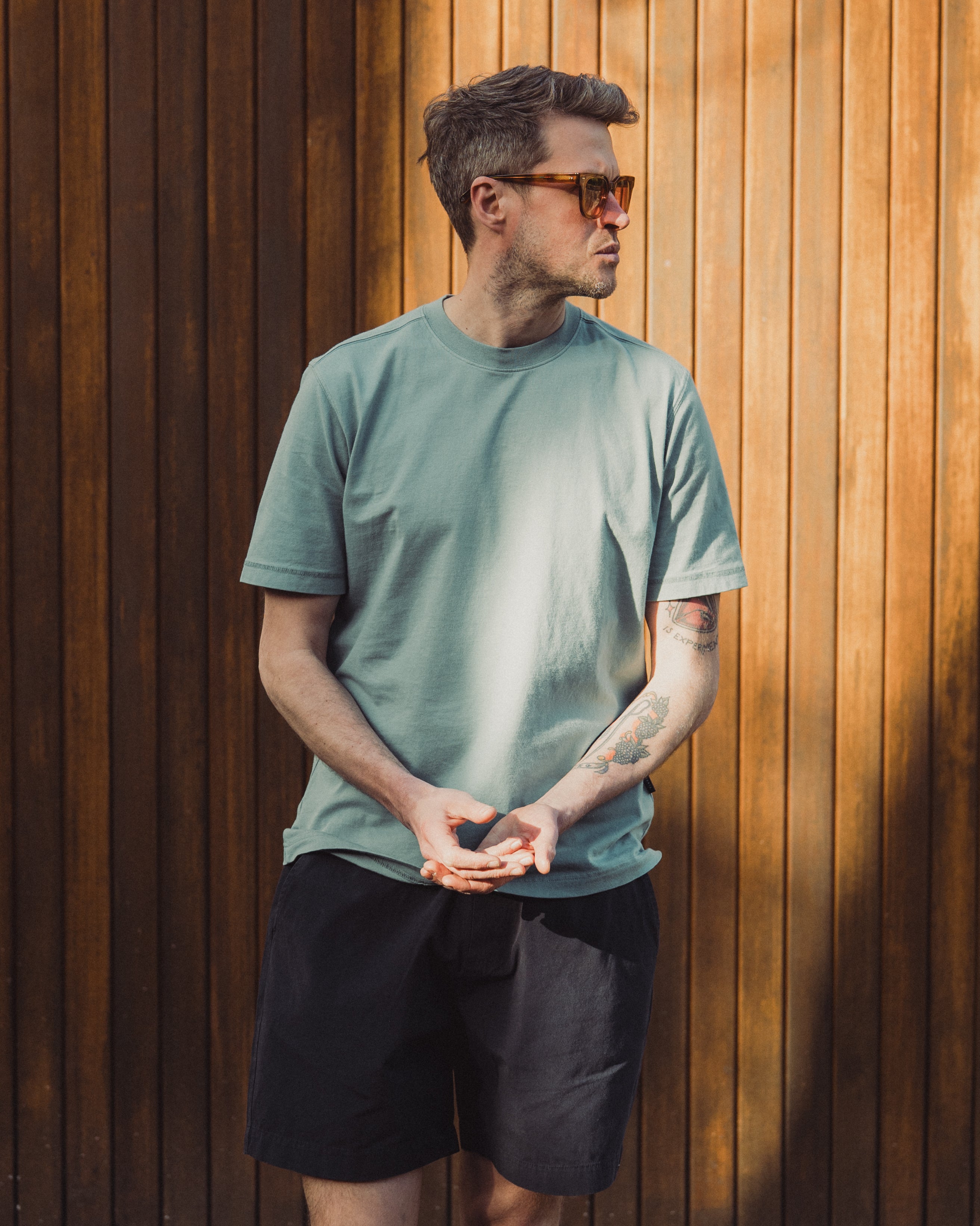 Man wearing a green t-shirt and black shorts standing against a wooden wall.
