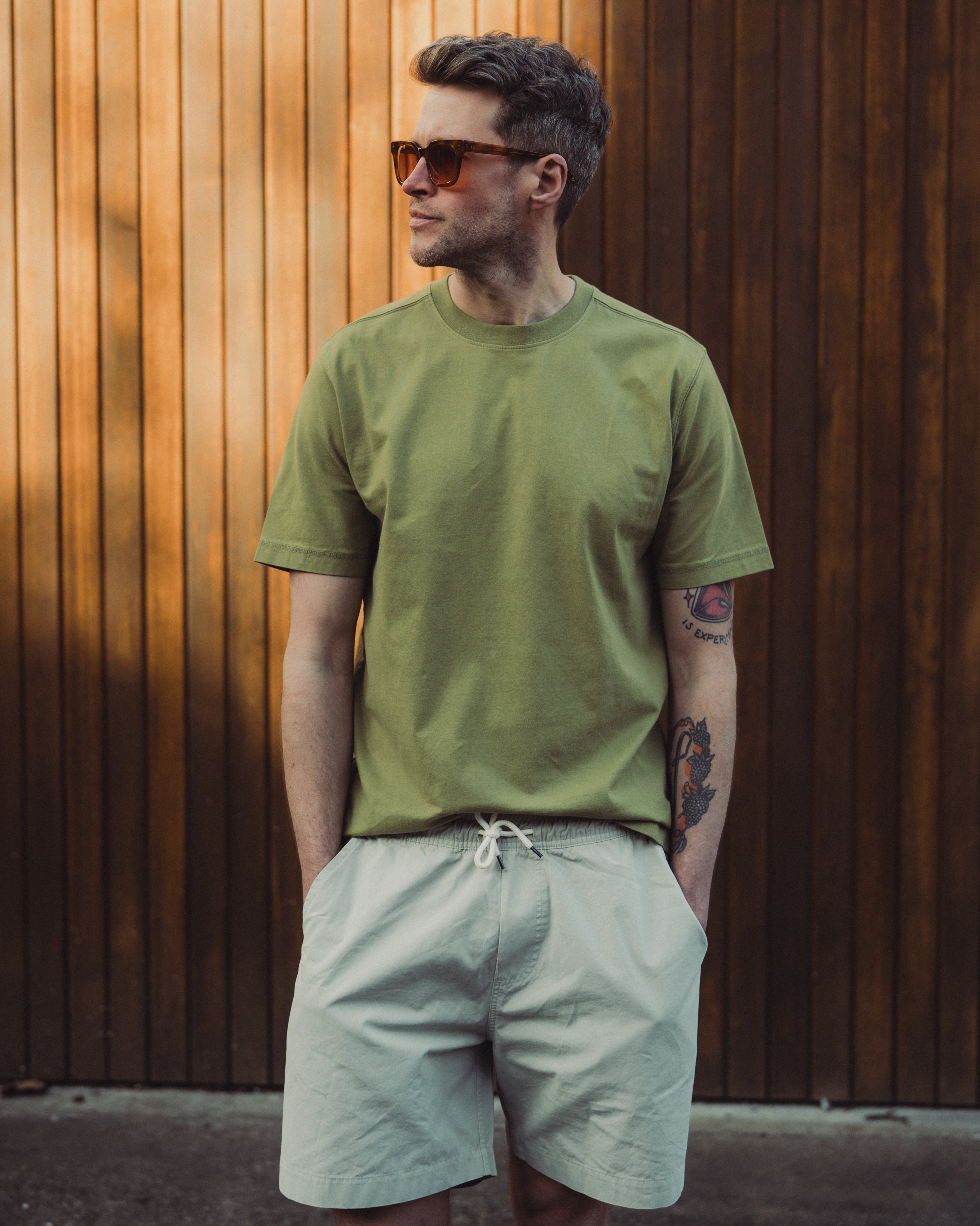 Man wearing a green t-shirt and light-colored shorts against a wooden wall.