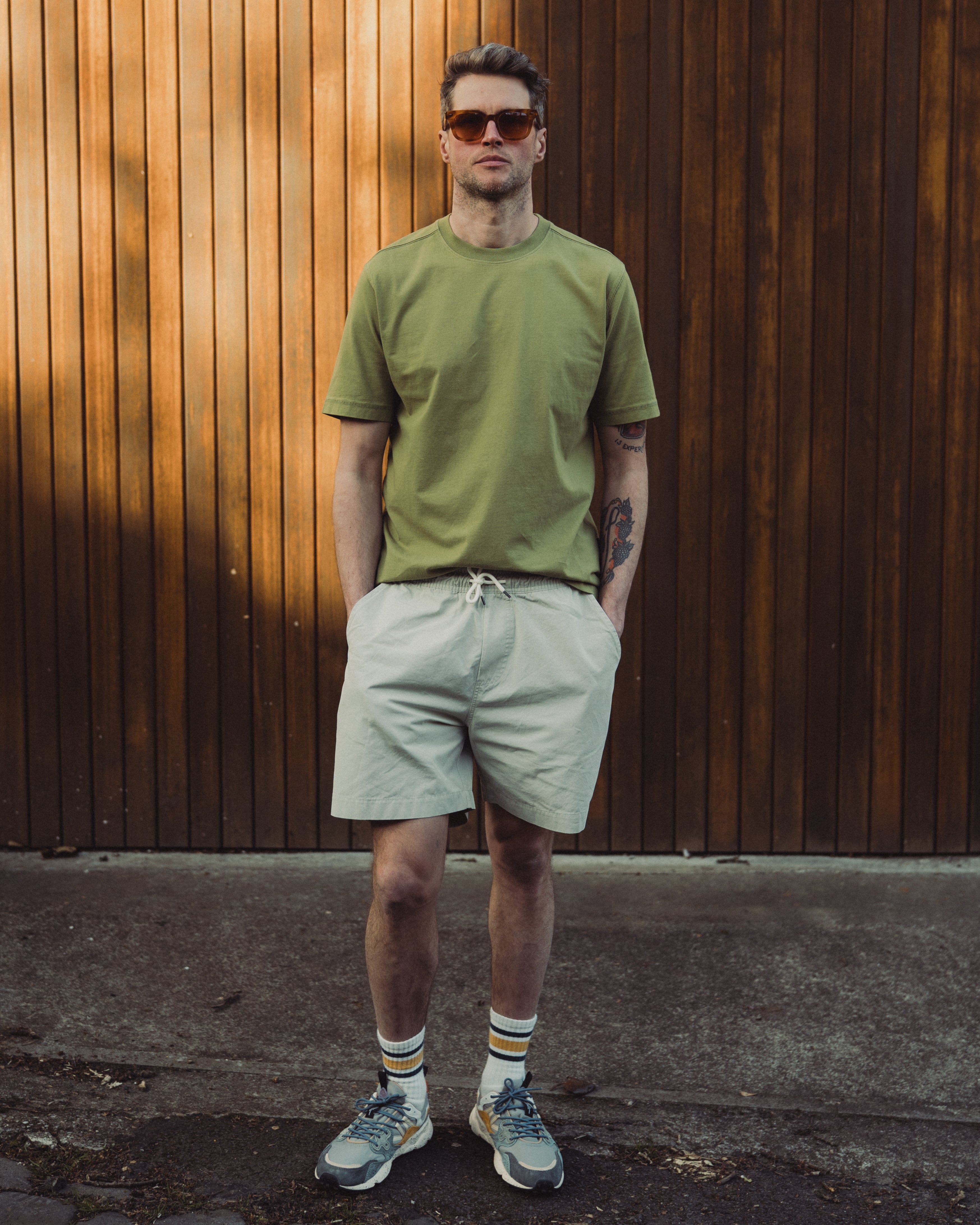 Man wearing a green t-shirt and light gray shorts standing against a wooden wall.
