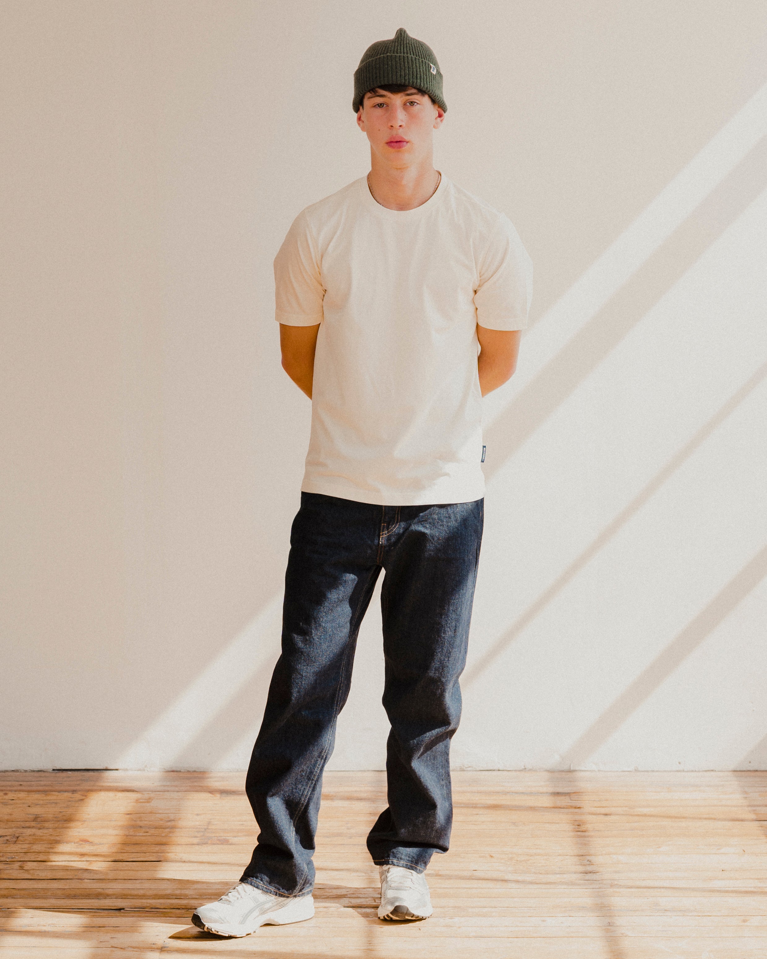 Person wearing a white t-shirt, dark jeans, and a cap standing in a room with light streaming through a window.