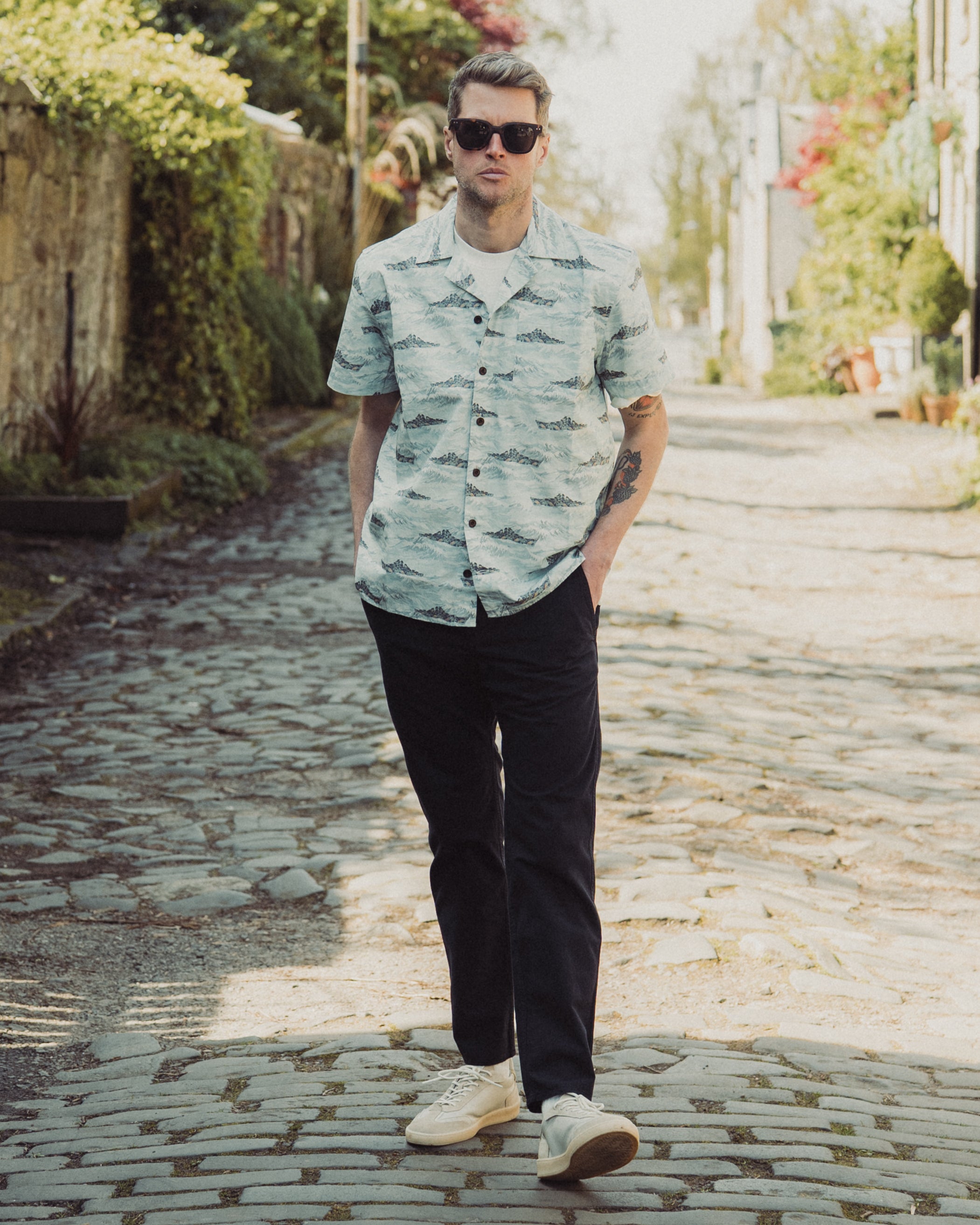 Man wearing a patterned shirt and sunglasses on a stone path with greenery.