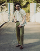 Man wearing sunglasses and a patterned shirt standing on a path with a white gate and greenery in the background.
